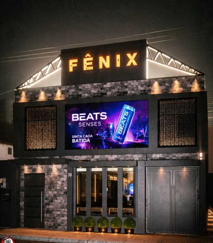 Fachada da Fênix Haus à noite, com o letreiro FÊNIX em lâmpadas e o telão de LED aceso